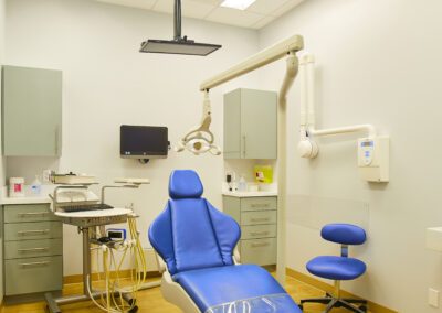 A bright dental examination room with a blue patient chair, dental equipment, and light-colored cabinets.