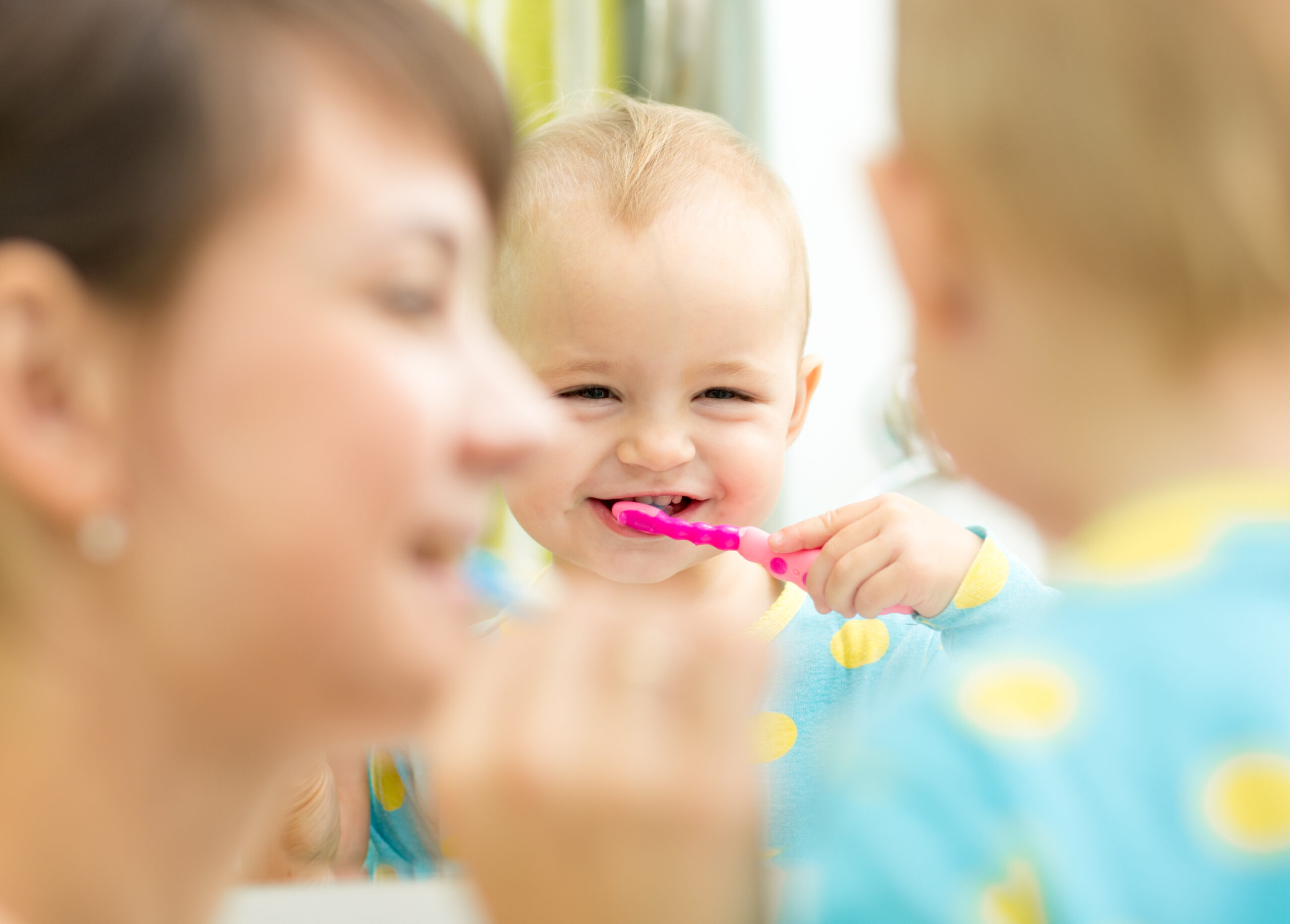 Smiling toddler in pajamas brushes teeth with a colorful toothbrush beside an adult, standing together at the bathroom mirror.